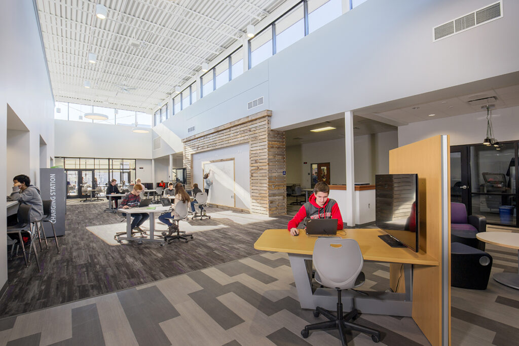 Students work on laptops and write at a whiteboard in collaboration spaces throughout an open, high-ceilinged hallway at DeForest Area High School