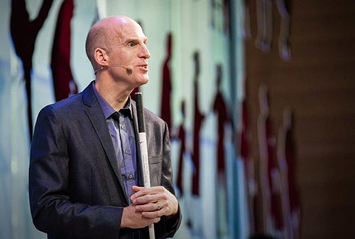 Chris Downey speaks in front of a large screen with a microphone while holding his white cane