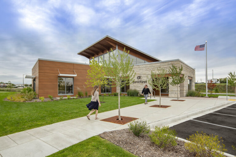 A treed sidewalk leads to a bank building accented with stone and wood