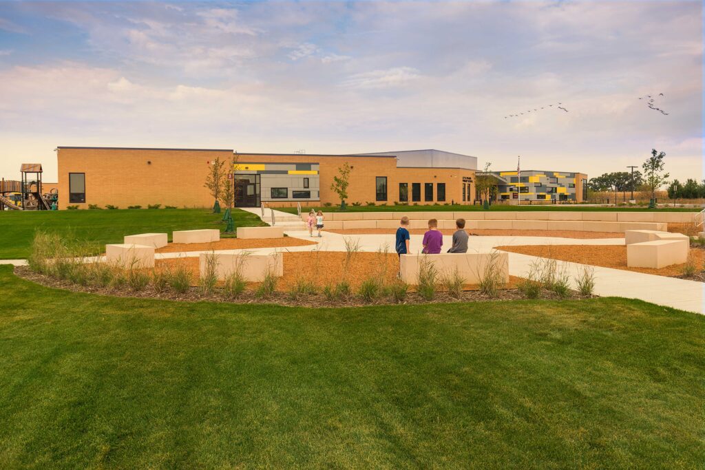 A exterior view of the outdoor classroom area with the school in the background. The outdoor classroom features large, rectangular boulders arranged in a ring around an intersection of walking paths.