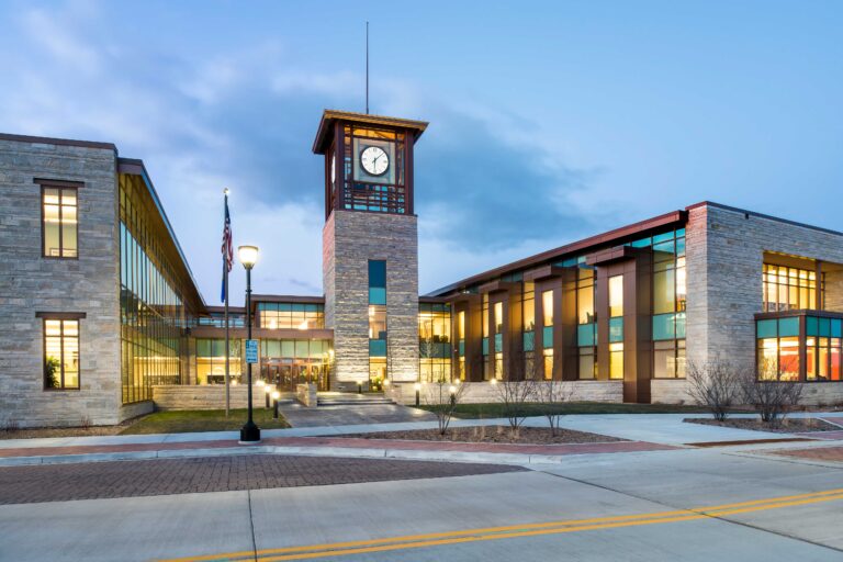 An exterior view at dusk of a U-shaped building with a tall clock tower in the middle at the Oak Creek City Hall and Public Library