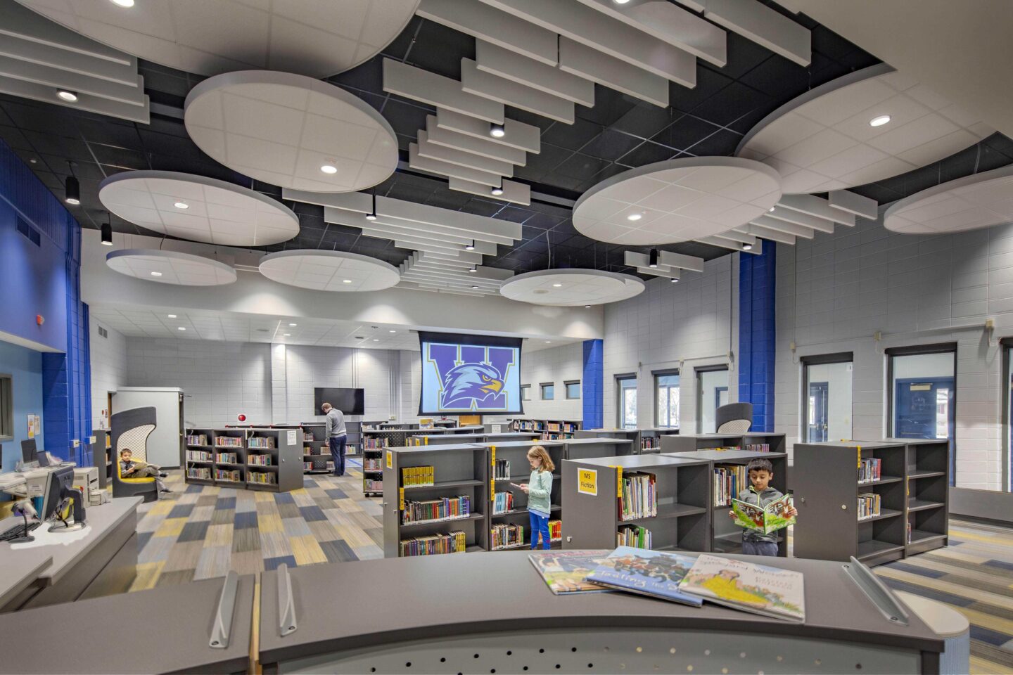 Geometric panels float high above a library with low shelves, as students browse books at Wheatland Center School