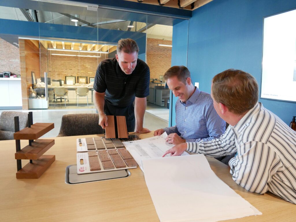 Three people meet around a table looking at product samples and plans, with additional glassed-in offices in the backgrounds, at the Bray Architects Milwaukee office
