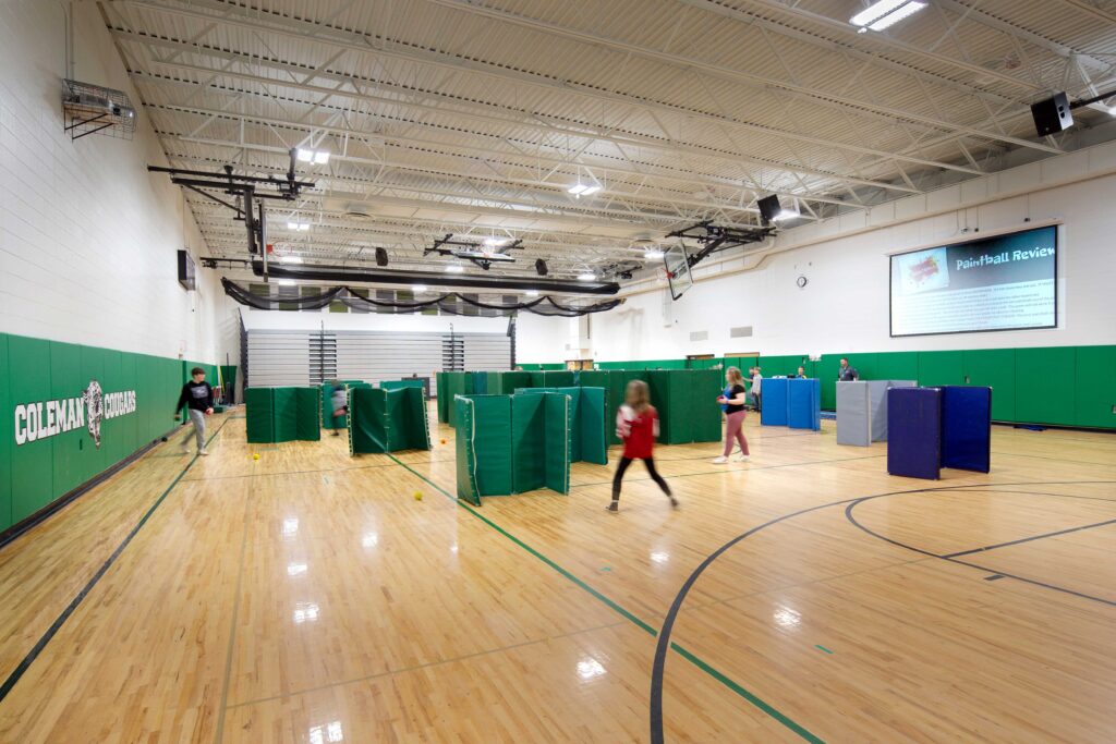 Bleaches, netting, and basketball goals at Coleman Elementary School can be retracted to create an open space, shown as students play around upright gym mats