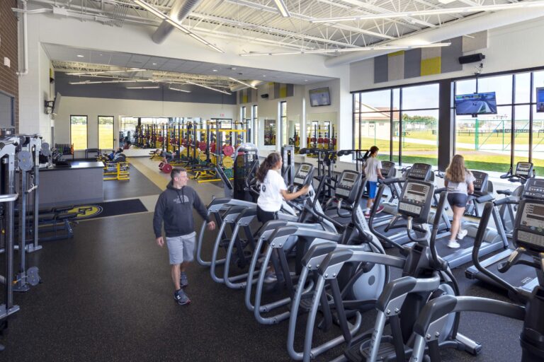 Students use elliptical trainers and other fitness equipment in a large, windowed athletics area