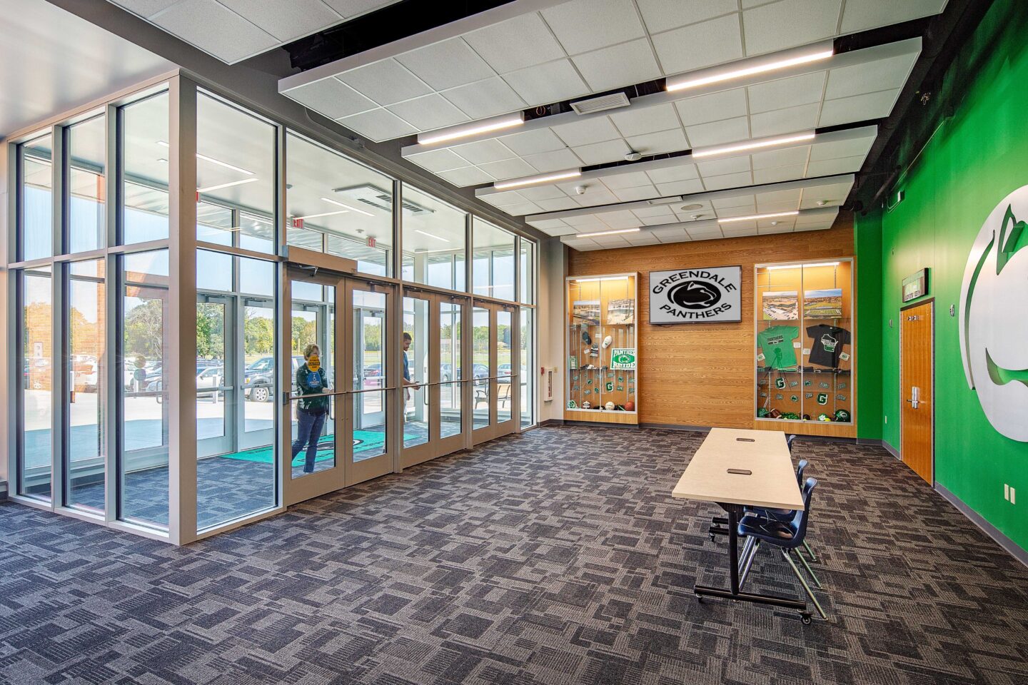 Community members come through a bright, glassed entrance to an athletics lobby accented by a trophy case and graphic wall