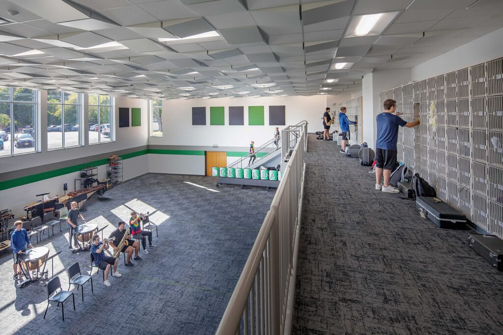 Students store instruments on a mezzanine that overlooks band students practicing in a large room, as natural light falls in from a set of windows