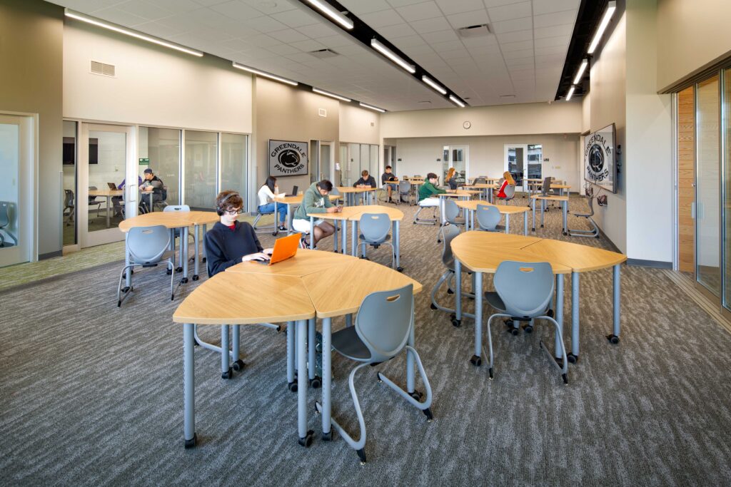 An open room with students working at tables is lined with glassed-in spaces for small-group work
