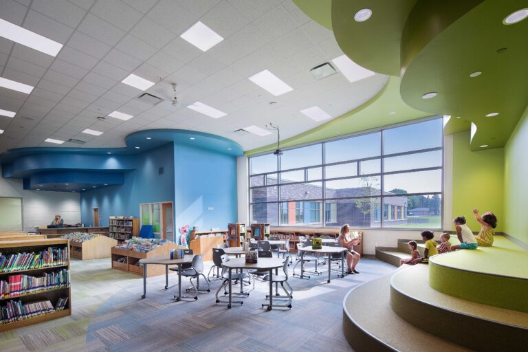 A teacher reads to students who are sitting on tiered steps in the school library, with a large window to the outdoors in the background