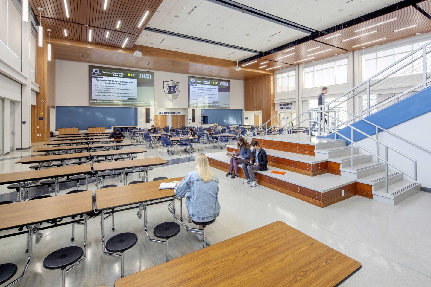 A modern learning staircase leads into a large, open forum with cafeteria-type seating and a wall of windows at Hudson High School