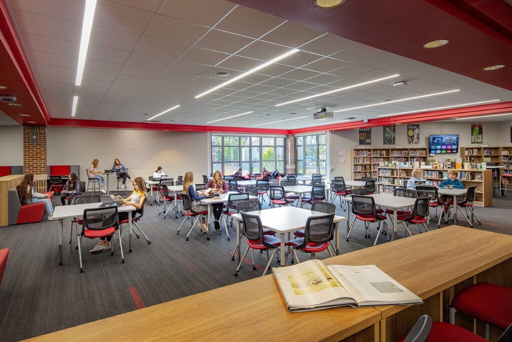 Students work at tables and seating areas in a media center featuring school colors and natural lighting at Les Paul Middle School