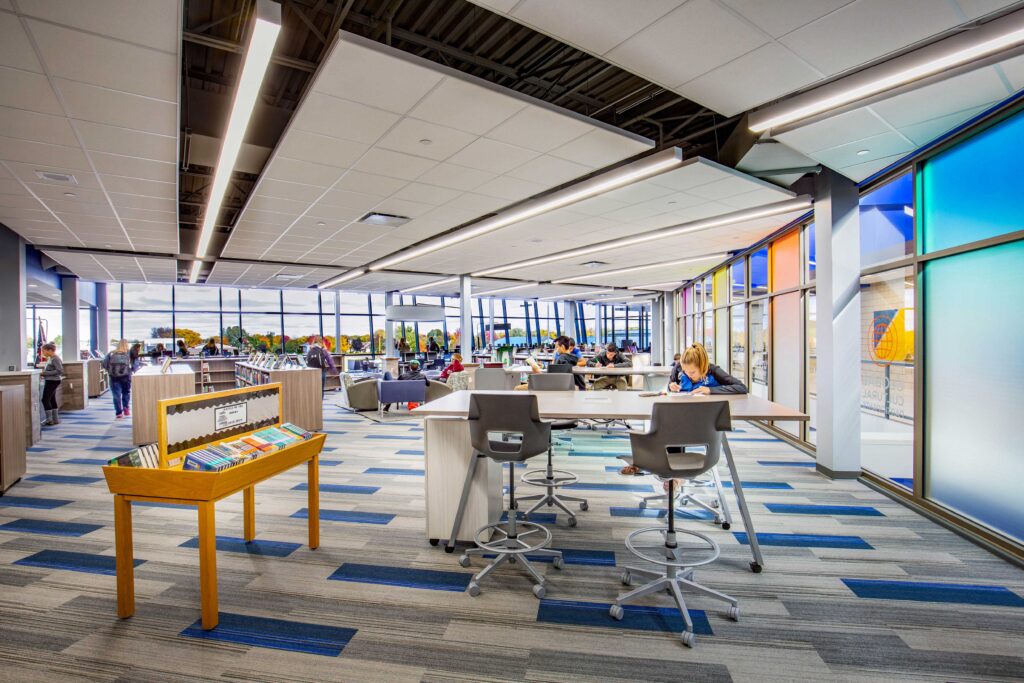 Windows surround a versatile library with low shelves, work tables, and collaboration areas at the Little Chute Intermediate, Middle, and High School