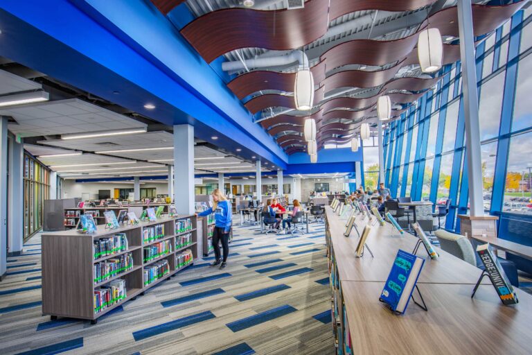 A library space at Little Chute Intermediate, Middle + High School features waves of wood on the ceiling connecting to a wall of windows around low shelves
