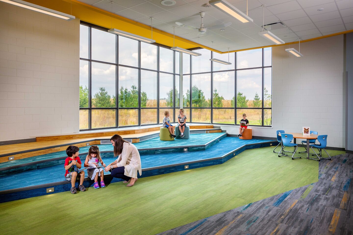 A teacher interacts with students on the tiered seating in the school library in front of large windows overlooking a natural landscape