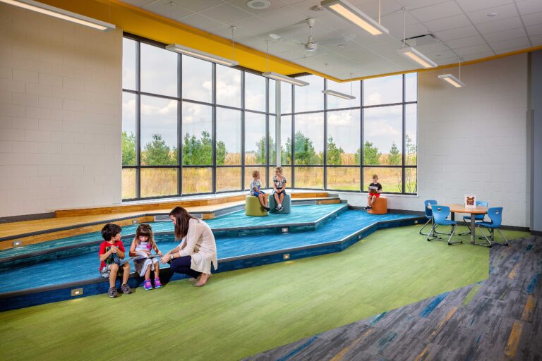 A teacher interacts with students on the tiered seating in the school library in front of large windows overlooking a natural landscape