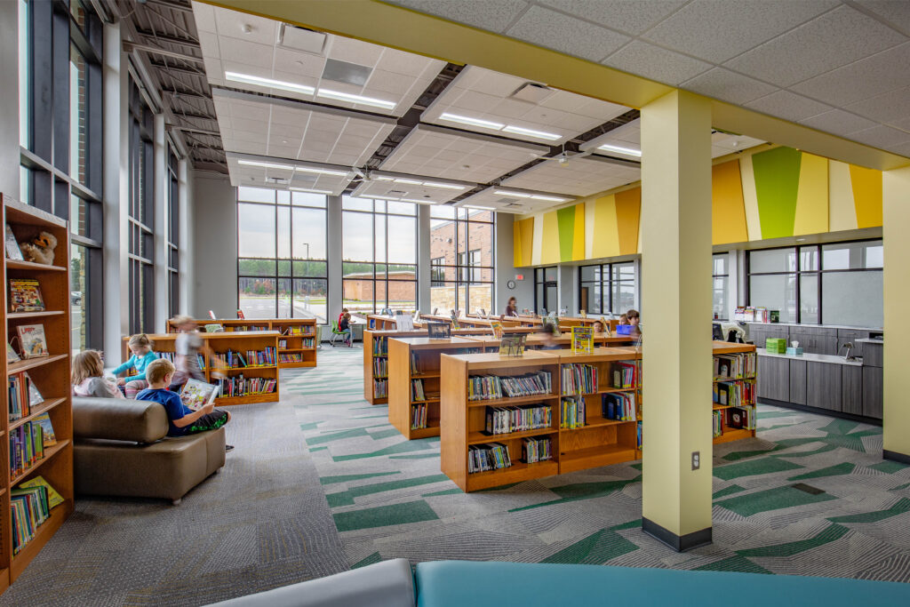 Two walls of windows let light flood into a library space with low shelves and colorful accents at Melrose-Mindoro School District