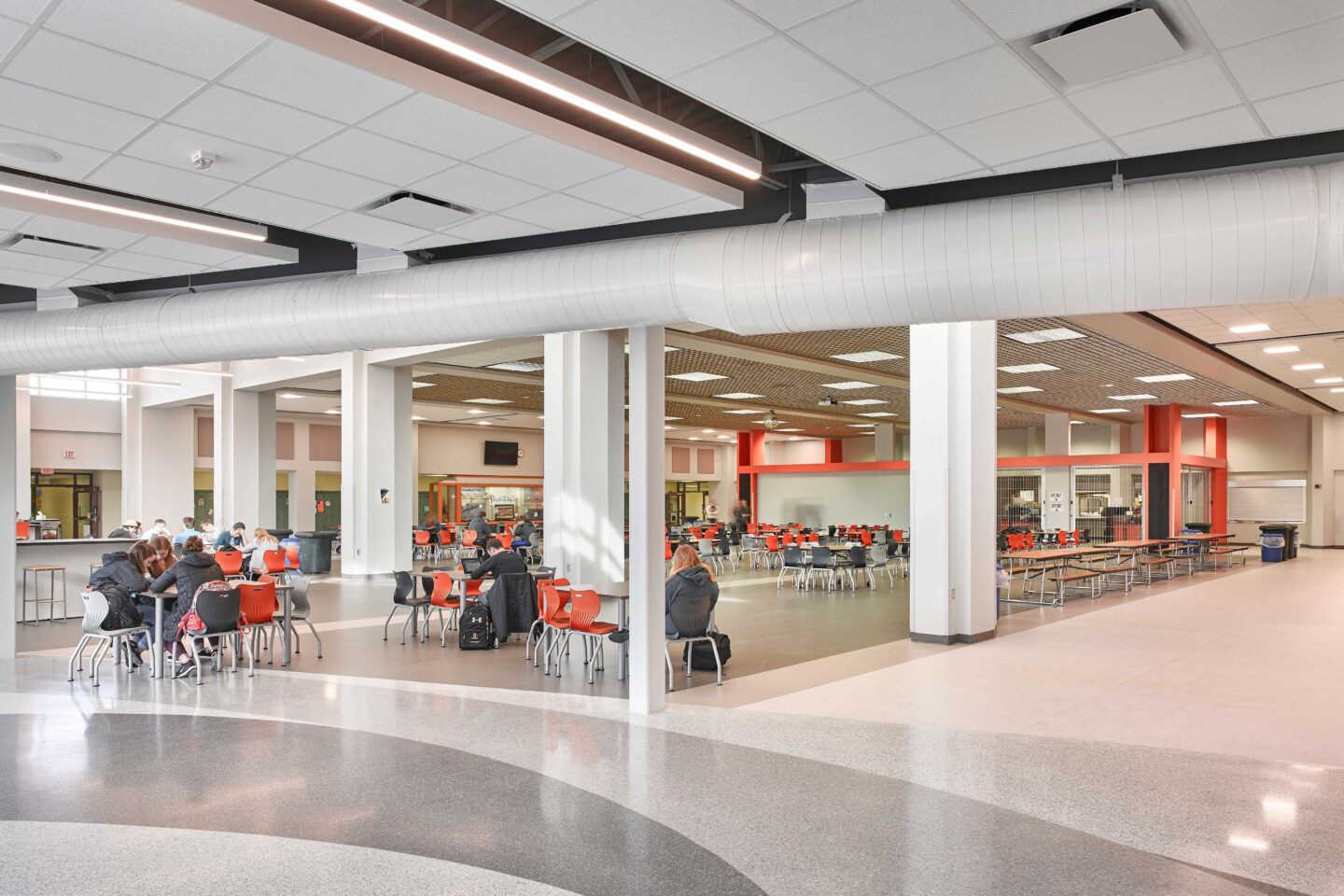 Students grouped around tables in the cafeteria, including a view of the kitchen area