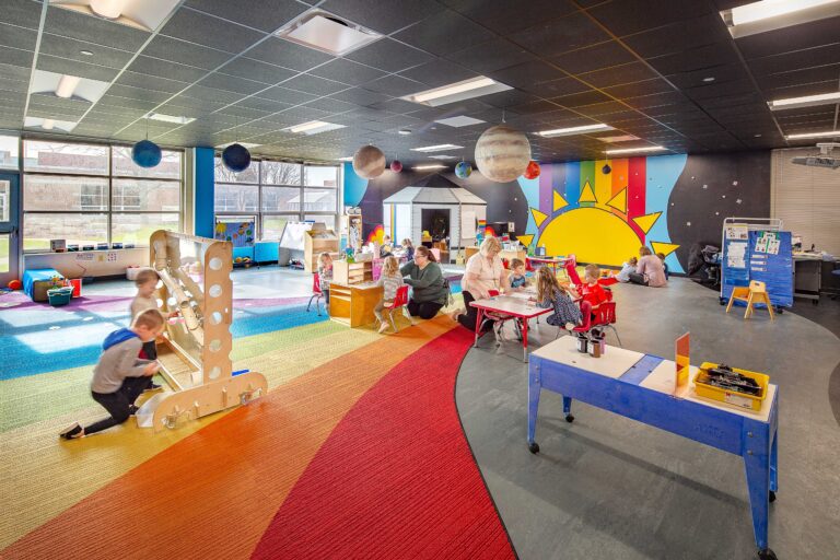 A rainbow wall mural floods into a rainbow carpet pattern as students work in a windowed classroom at Pewaukee Lake Elementary School