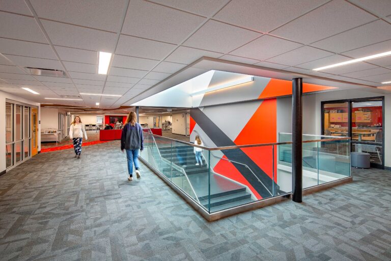 Students walk through a landing area above a modern, glassed-in staircase with red accents at Pewaukee High School