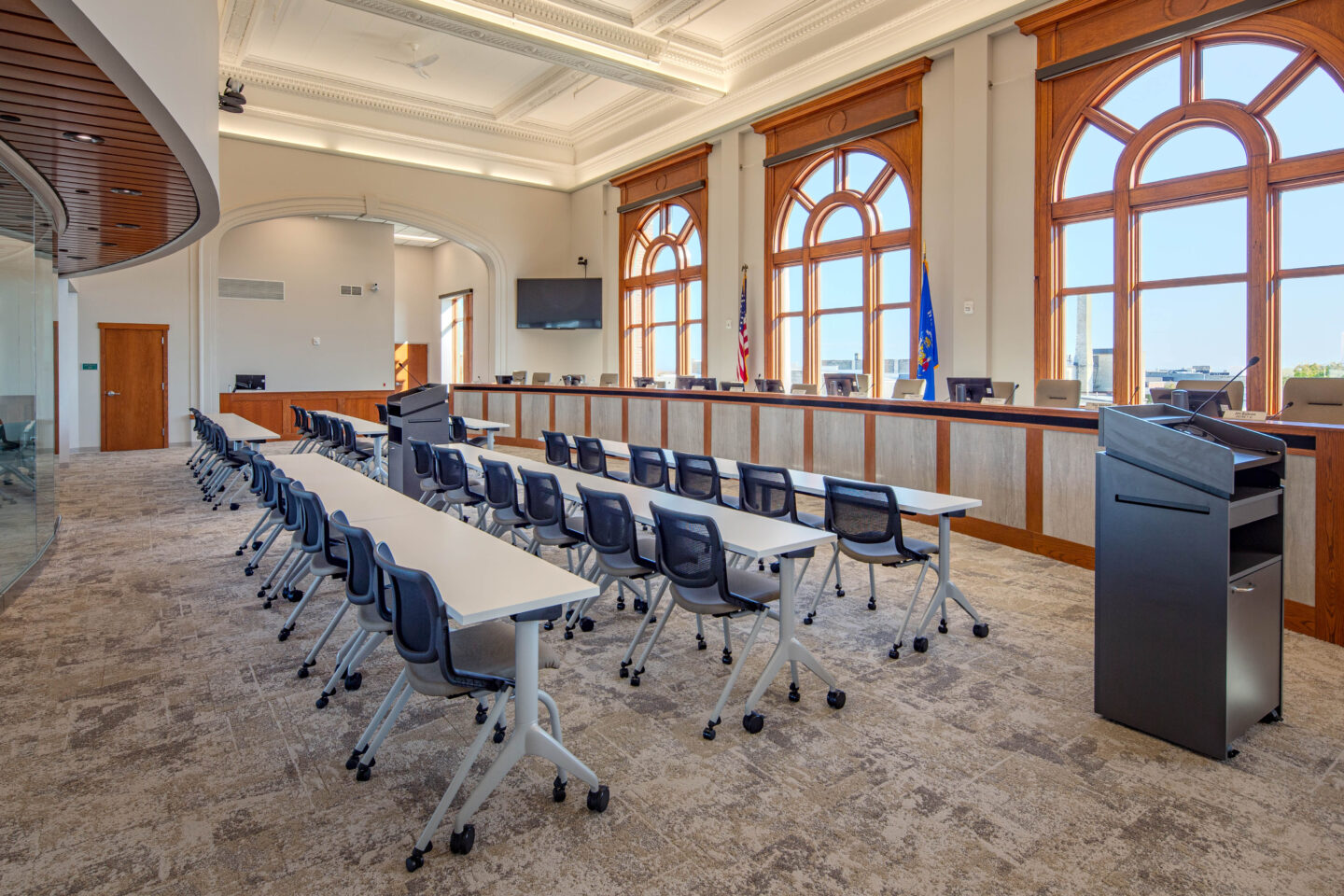 A dais faces rows of tables and chairs, backed by arched windows and topped off with a stained glass skylight, in the council chamber at Sheboygan City Hall