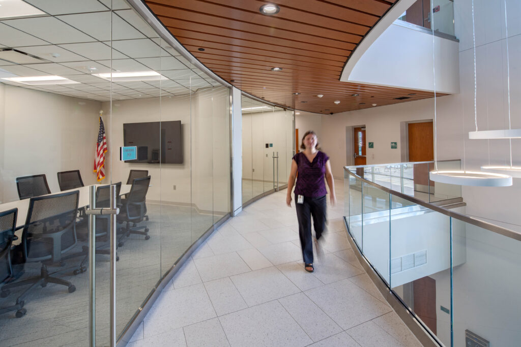 A person walks down a curved hallway that overlooks an atrium, with glassed-in conference rooms to one side