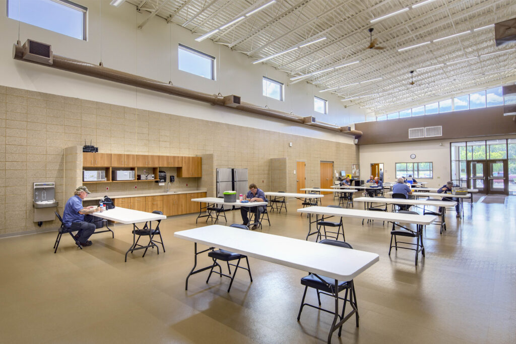 Staff members sit at tables throughout an open commons area, with windows and glass doors providing natural light