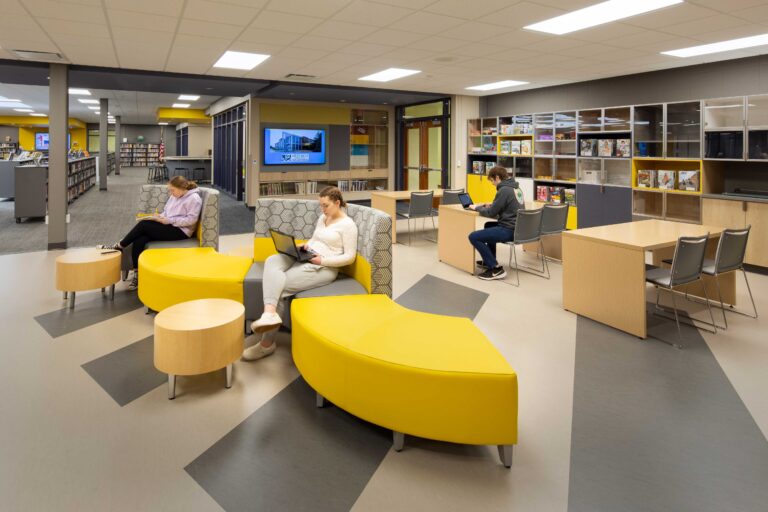 Students study at colorful, flexible furniture arranged through a seating space with a library in the background