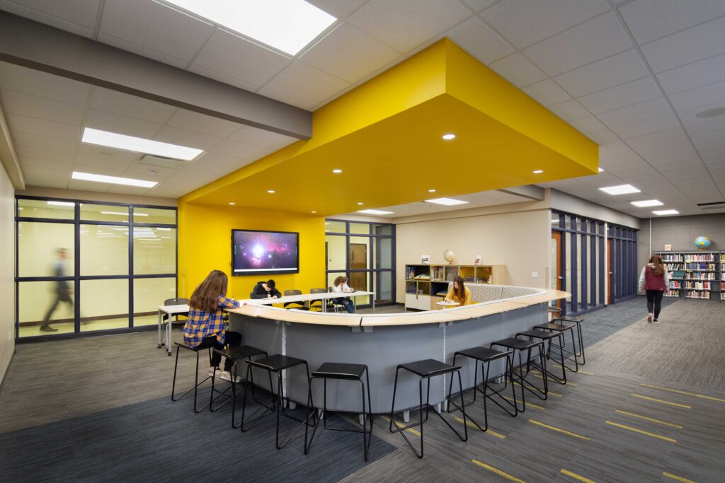 A counter curves around a study space accented by a yellow ceiling call-out with windows that connect to an adjacent hallway