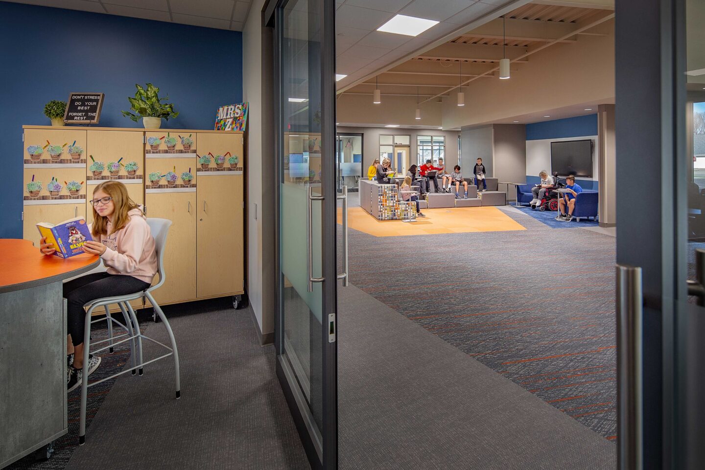 A student reads at a desk in a classroom while other students collaborate at one of the nearby resource areas visible through large sliding glass doors