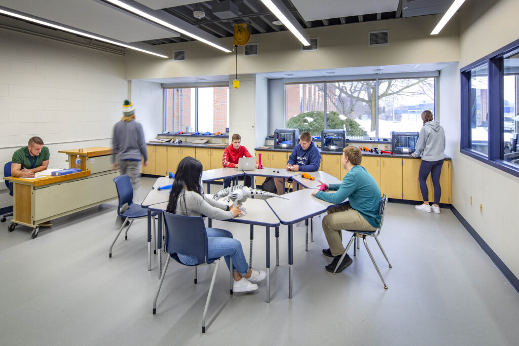 Students gather around a table and countertop work areas in a window-lined classroom at Whitnall High School