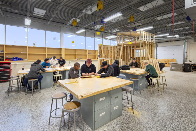 Students group around tall tables with a wooden structure under construction at a woods shop in Whitnall High School