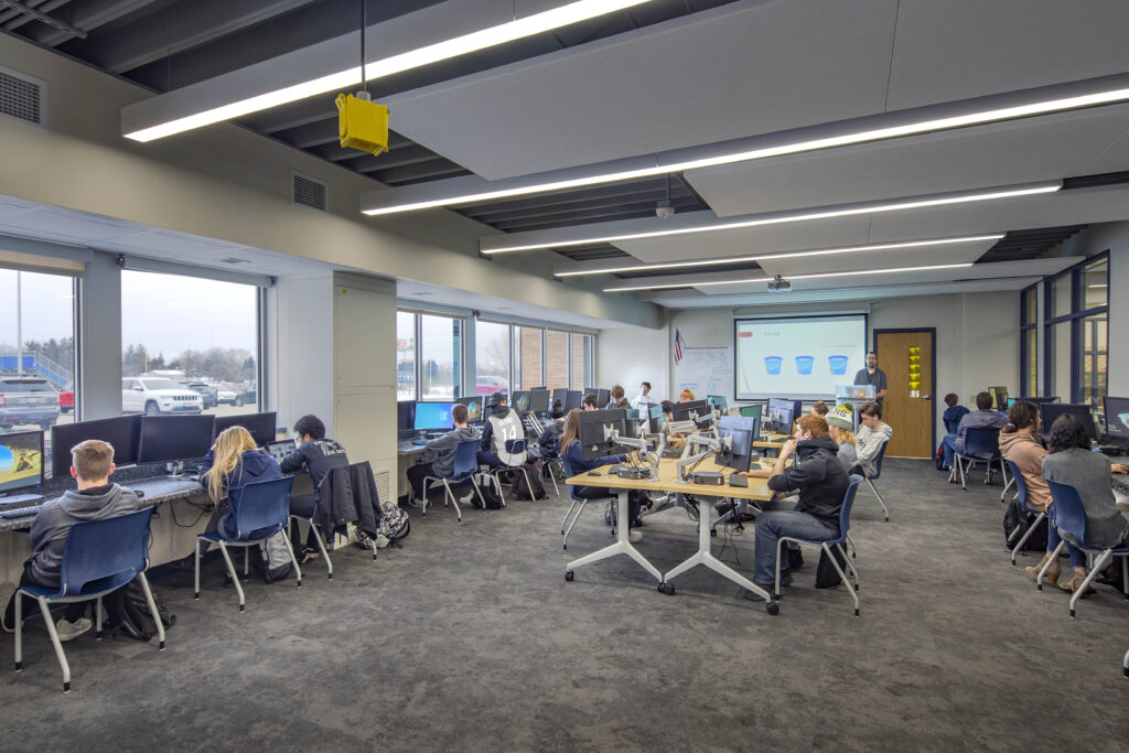 A large, windowed classroom with tables and computer stations is filled with students at work in Whitnall High School