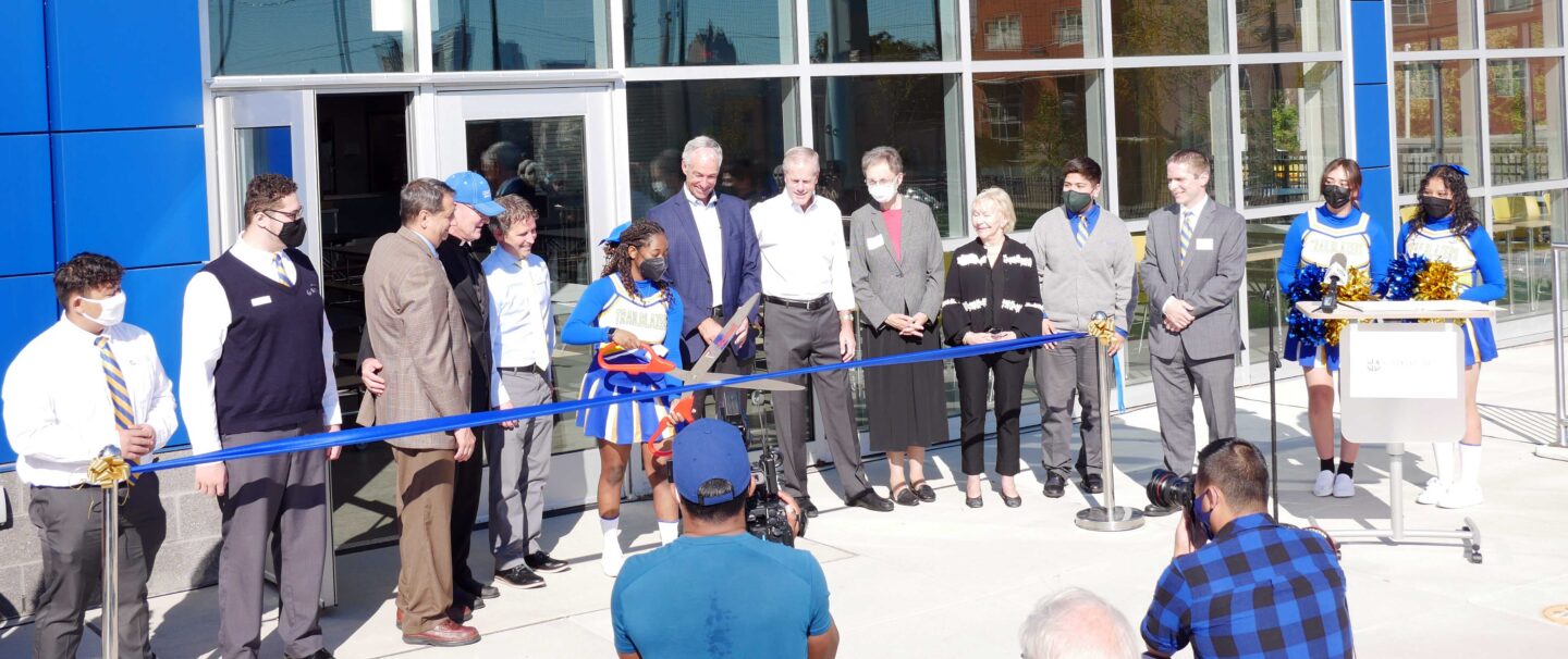 A row of people stand behind a ribbon on two stanchions as a student with oversized scissors prepares to cut the ribbon at Cristo Rey High School in Milwaukee