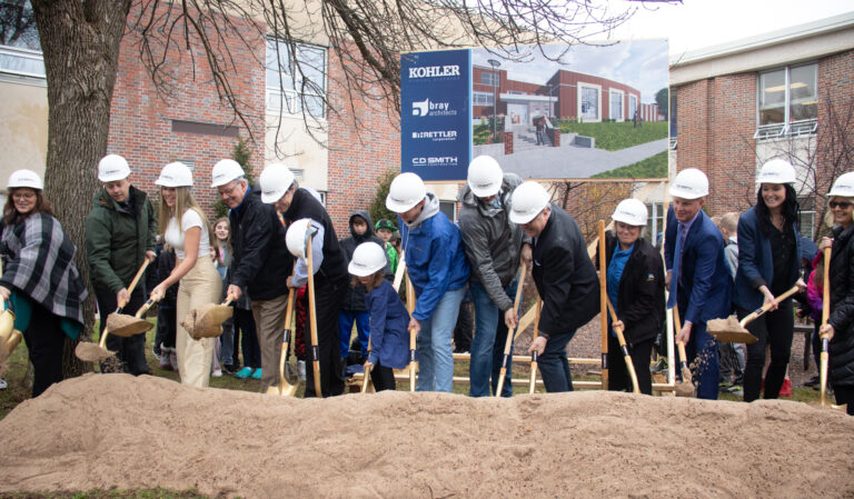 A row of people in hardhats dig shovels into the ground for a ceremonial groundbreaking at the Kohler School District