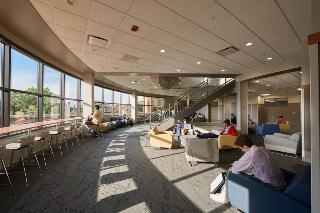 Students study in chairs and high-top seating in front of a curved bay of windows in the lower lounge