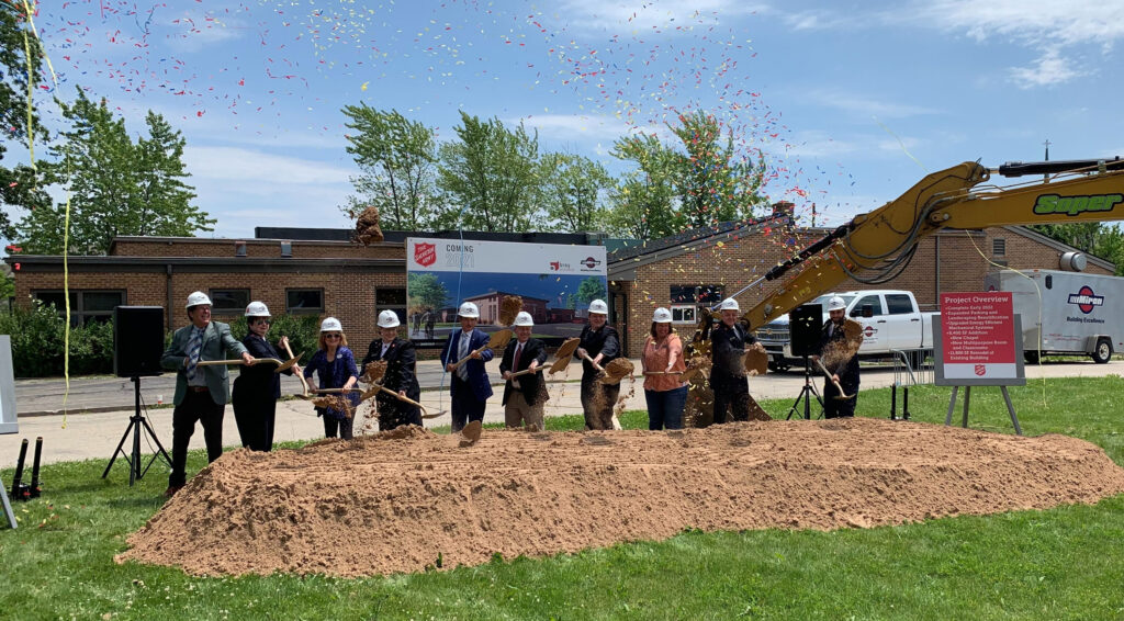 A row of people in hard hats dig from a pile of dirt as confetti showers them